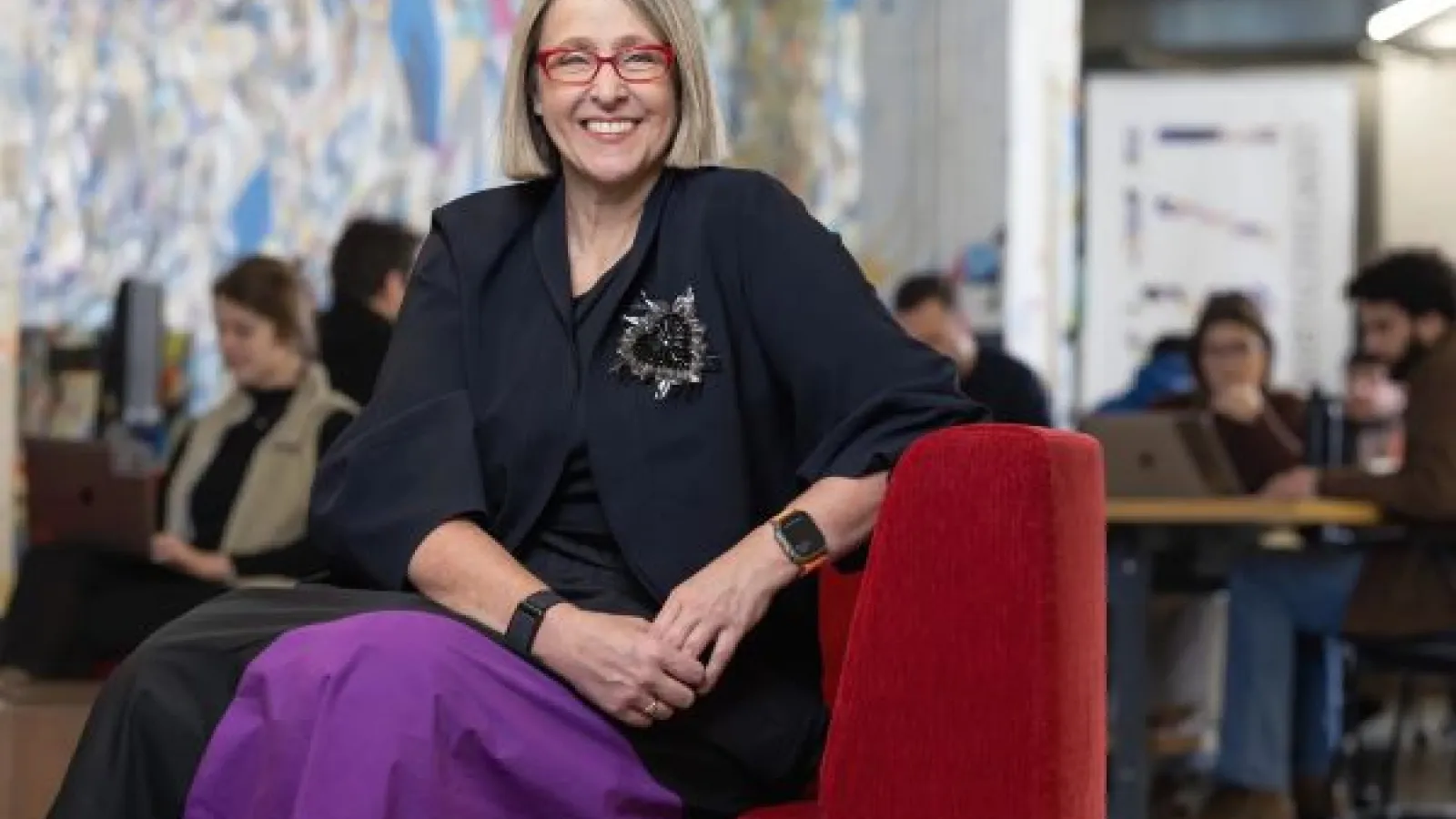 a woman sits in a red velvet chair, smiling at the camera